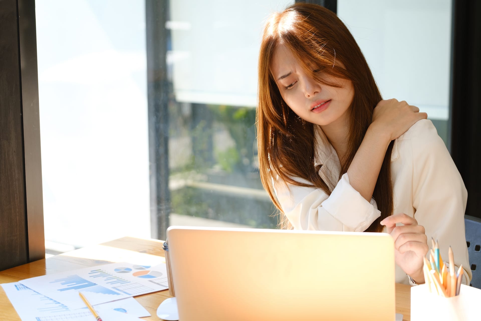 A young lady sitting at a computer holding her upper back. She has long dark hair and is wearing a white shirt.